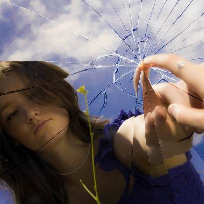 Portrait of Sam McMeeking looking into a broken mirror. Here finger is touching the middle of the crack. The reflection of the mirror shows a yellow flower and a blue sky.