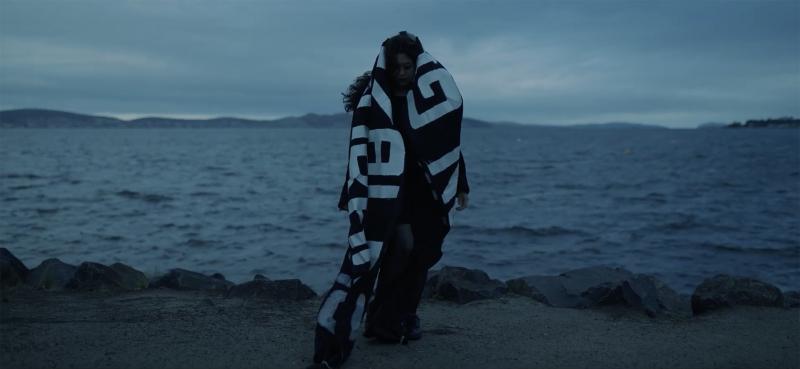 Still from Baltimöre Charlót's music video Claws. Baltimöre Charlót is standing on a beach wrapped in a black blanket with white text. The image is dark and moody.